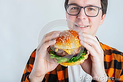 A young guy with glasses holding a fresh Burger. A very hungry student eats fast food. Hot helpful food. The concept of gluttony a Stock Photo