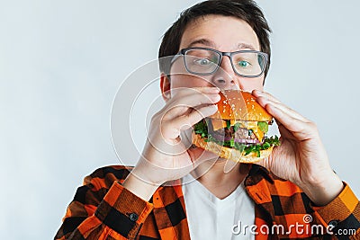 A young guy with glasses holding a fresh Burger. A very hungry student eats fast food. Hot helpful food. The concept of gluttony a Stock Photo