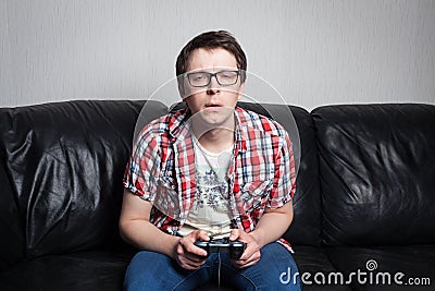 Young guy with glasses and red shirt playing video games on the joystick, sitting on a black leather sofa Stock Photo