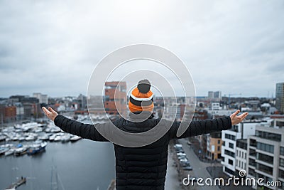 A young guy look at city from the roof of building with his arms raised. Black jacket and orange cap. Belgium, Antwerp. Stock Photo