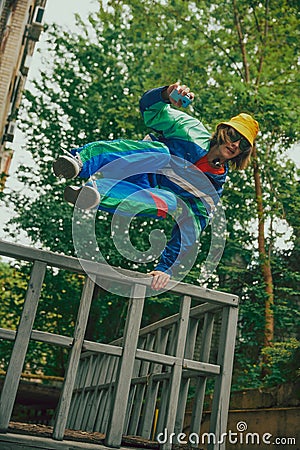 Young guy in 90s style tracksuit and yellow panama, with retro camera, having fun outdoor, jumping over the railing Stock Photo