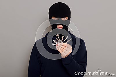 A young guy thief holds stolen keys from house or other property in his hands, wearing hoodie and robber mask, looks at camera, Stock Photo