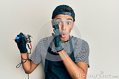 Young handsome african american man tattoo artist wearing professional uniform and gloves holding tattooer machine hand on mouth Stock Photo