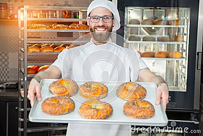 A young handsome baker holding fresh bagels with poppy seeds on a tray on the background of an oven and a rack with baked goods. Stock Photo