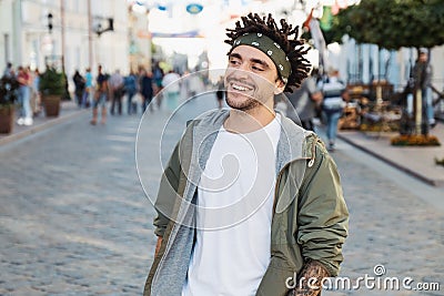 Young handsome beard man dressed stylish casual clothes, white t-shirt, green jacket, bandana posing outdoor. Smiling hipster with Stock Photo