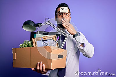 Young handsome businessman with beard holding cardboard box with fired reminder on head cover mouth with hand shocked with shame Stock Photo