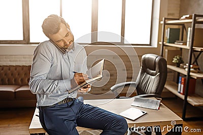 Young handsome businessman sit on table and talk on phone in his own office. He write in notebook. Business call Stock Photo