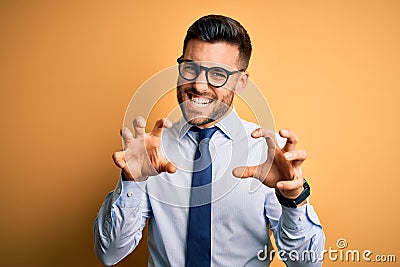 Young handsome businessman wearing tie and glasses standing over yellow background smiling funny doing claw gesture as cat, Stock Photo