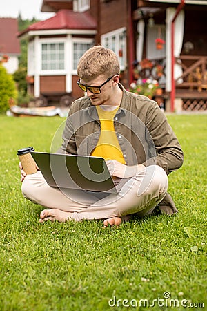 Young handsome businessman works on laptop outdoors, sits with coffee on grass Stock Photo
