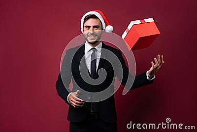 Young handsome caucasian guy in business suit and Santa hats stands on red background in studio and smilie throws up red gift box Stock Photo
