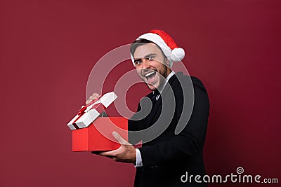 Young handsome caucasian guy in business suit and Santa hats stands on red background in studio and teeth smilie Holding red gift Stock Photo