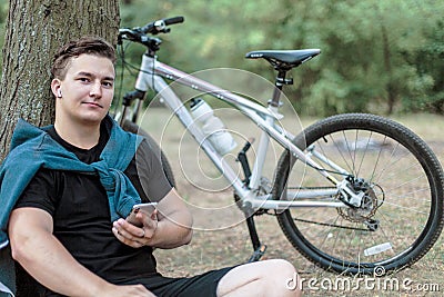 Young handsome caucasian man slightly smiling, holds mobile phone, sitting on the ground close to the big tree. Stock Photo