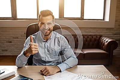 Young handsome cheerful businessman sit on table and pose in his own office. He hold big thumb up and smile. Nice happy Stock Photo