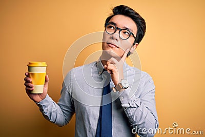 Young handsome chinese man drinking glass of take away coffee over yellow background serious face thinking about question, very Stock Photo