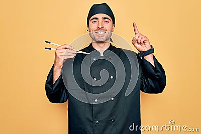 Young handsome cooker man with blue eyes wearing uniform and hat using chopsticks surprised with an idea or question pointing Stock Photo