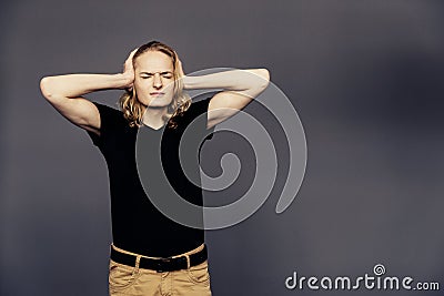 Young handsome guy in a black T-shirt with long blonde hair covered his ears with his hands from annoying noise on a gray Stock Photo