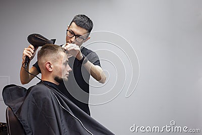 Handsome guy has a haircut in a barbershop, a Kazakh barber dries a client`s hair dryer against a gray wall, a hairdresser makes Stock Photo