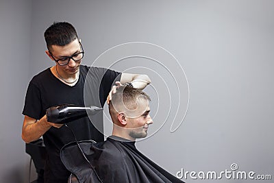 Handsome guy has a haircut in a barbershop, a Kazakh barber dries a client`s hair dryer against a gray wall, a hairdresser makes Stock Photo