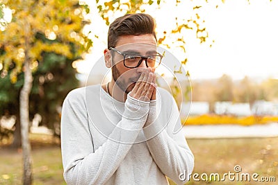 Young handsome hipster man in glasses and a white sweater is walking in a cold autumn park. Heats his hands with his breath. Stock Photo