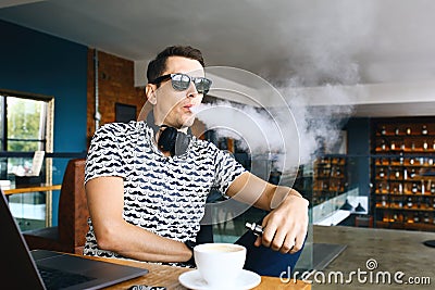 Young handsome hipster man insunglasse sitting in cafe with a cup of coffee, vaping and releases a cloud of vapor Stock Photo