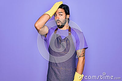 Young handsome hispanic clenaer man wearing housework apron and washing gloves surprised with hand on head for mistake, remember Stock Photo