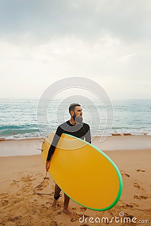 Young handsome male surfer with a surfboard is enjoying a view while walking a sandy beach at sea. Summer, vacation, ocean. Stock Photo