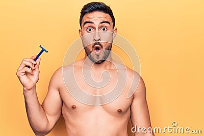 Young handsome man with beard shirtless holding depilation razor over yellow background scared and amazed with open mouth for Stock Photo