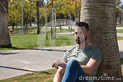 Young and handsome man, with blue eyes, beard, green t-shirt and jeans, sitting and leaning on a palm tree trunk and with a Stock Photo