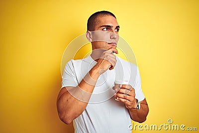 Young handsome man drinking a take away glass of coffee over yellow isolated background serious face thinking about question, very Stock Photo