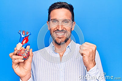 Young handsome man holding heart organ with veins and arteries over blule background screaming proud, celebrating victory and Stock Photo