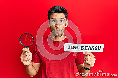 Young handsome man holding magnifying glass for job search making fish face with mouth and squinting eyes, crazy and comical Stock Photo