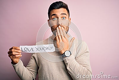 Young handsome man holding paper with self love message over pink background cover mouth with hand shocked with shame for mistake, Stock Photo