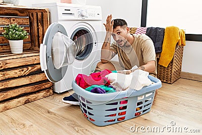 Young handsome man putting dirty laundry into washing machine surprised with hand on head for mistake, remember error Stock Photo