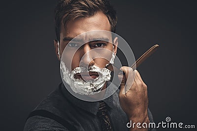 Young handsome man with shaving cream on his face, grooming his beard with straight razor and looking Stock Photo