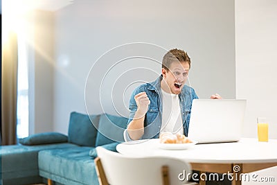 Young handsome man after successful online negotiations working in his latop from homewhile having croissant and fresh Stock Photo