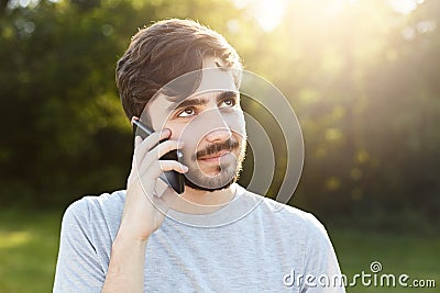 Young handsome man with thick beard and dark big eyes holding smart phone phoning his friend while standing at green background. H Stock Photo