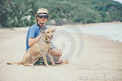 Young handsome man wearing blue t-shirt, hat and sunglasses, sitting on the beach with the dog in Thailand Stock Photo