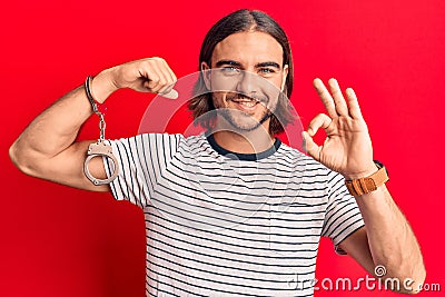 Young handsome man wearing prisoner handcuffs doing ok sign with fingers, smiling friendly gesturing excellent symbol Stock Photo