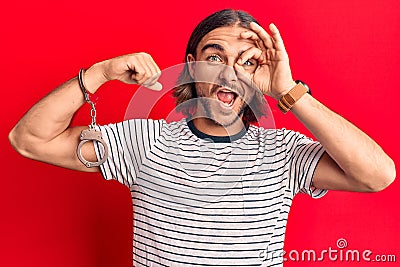 Young handsome man wearing prisoner handcuffs smiling happy doing ok sign with hand on eye looking through fingers Stock Photo