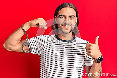 Young handsome man wearing prisoner handcuffs smiling happy and positive, thumb up doing excellent and approval sign Stock Photo