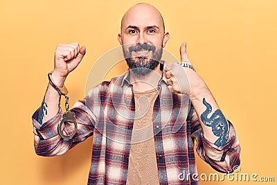 Young handsome man wearing prisoner handcuffs smiling happy and positive, thumb up doing excellent and approval sign Stock Photo