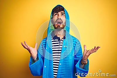 Young handsome man wearing rain coat with hood standing over isolated yellow background crazy and mad shouting and yelling with Stock Photo