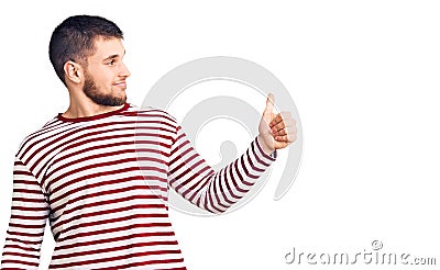 Young handsome man wearing striped sweater looking proud, smiling doing thumbs up gesture to the side Stock Photo