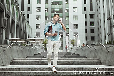Young handsome office worker talking on smartphone holding laptop and cup of coffee walking down the stairs at business Stock Photo