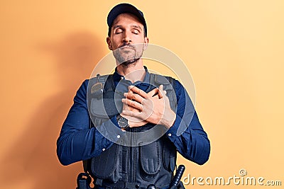 Young handsome policeman wearing police uniform and bulletprof over yellow background smiling with hands on chest, eyes closed Stock Photo