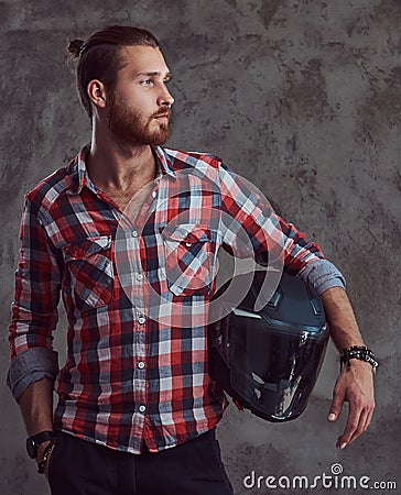 Young handsome redhead biker in a flannel shirt, holds motorcycle helmet, posing on a gray background. Stock Photo