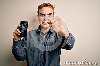 Young handsome redhead man holding broken smartphone showing cracked screen annoyed and frustrated shouting with anger, yelling Stock Photo