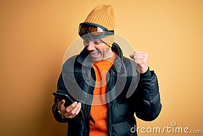 Young handsome skier man wearing ski goggles using smartphone over yellow background screaming proud and celebrating victory and Stock Photo