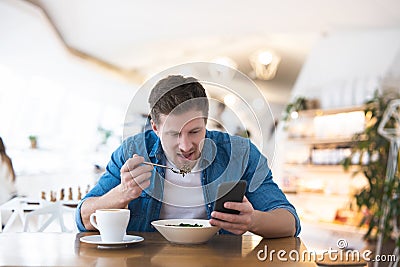 Young handsome smiling man drinks his hot coffee and eats salad for lunch while reading message in his smartphone during break at Stock Photo