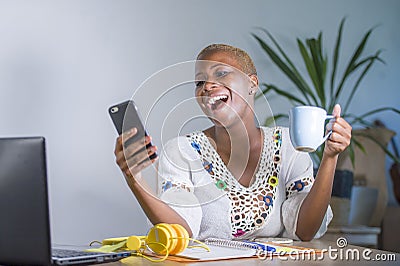 Young happy and attractive black afro american hipster woman working at home office with laptop computer using internet on mobile Stock Photo